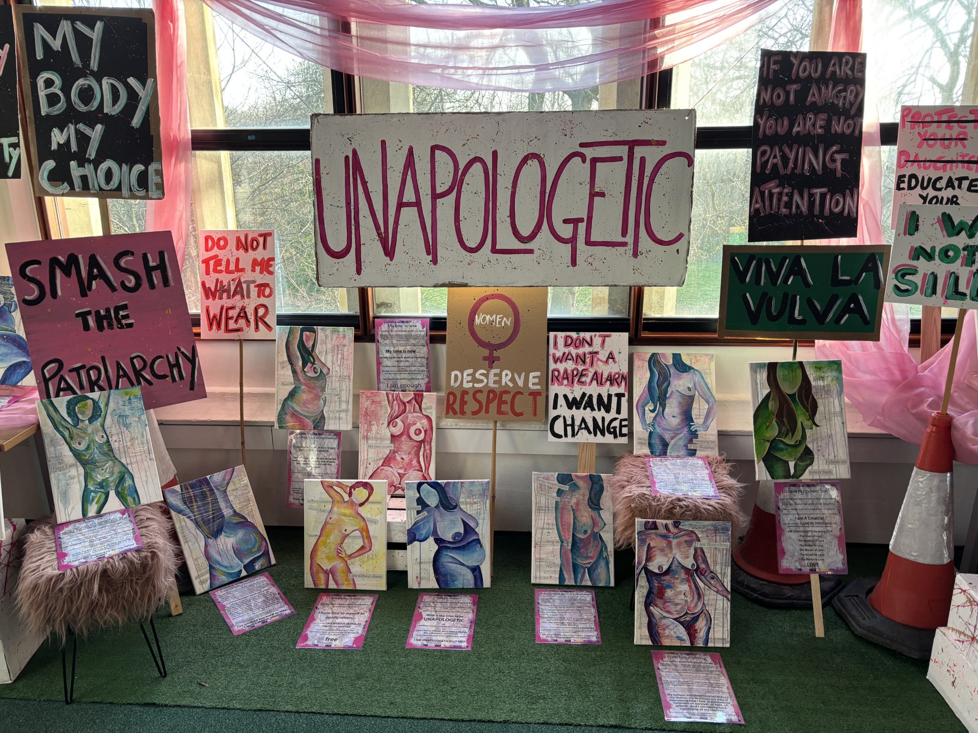 “Women’s rights art installation featuring protest signs reading ‘Unapologetic,’ ‘My Body My Choice,’ ‘Smash the Patriarchy,’ and body-positive paintings of diverse female forms arranged in a powerful feminist display.”
