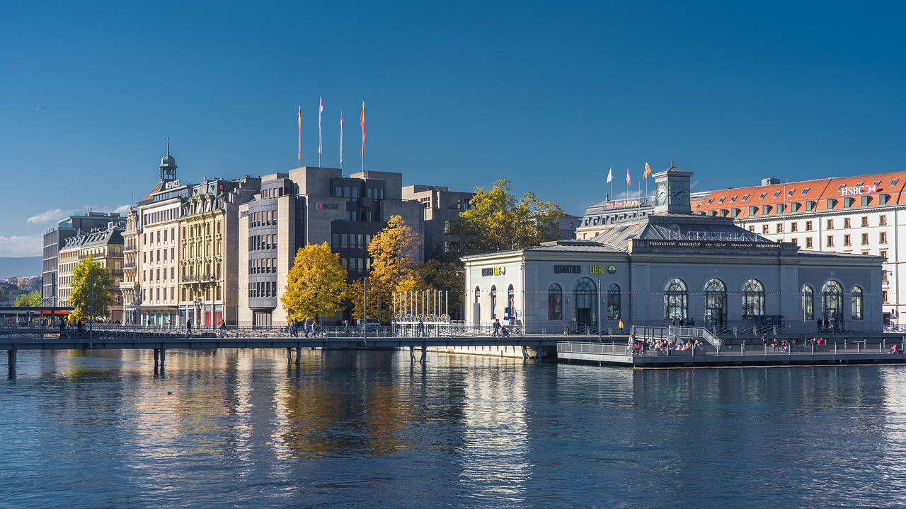 A panoramic view of Geneva, Switzerland, featuring the city’s lakeside architecture, a bridge crossing Lake Geneva, flowing water, and a backdrop of natural landscapes and mountains.