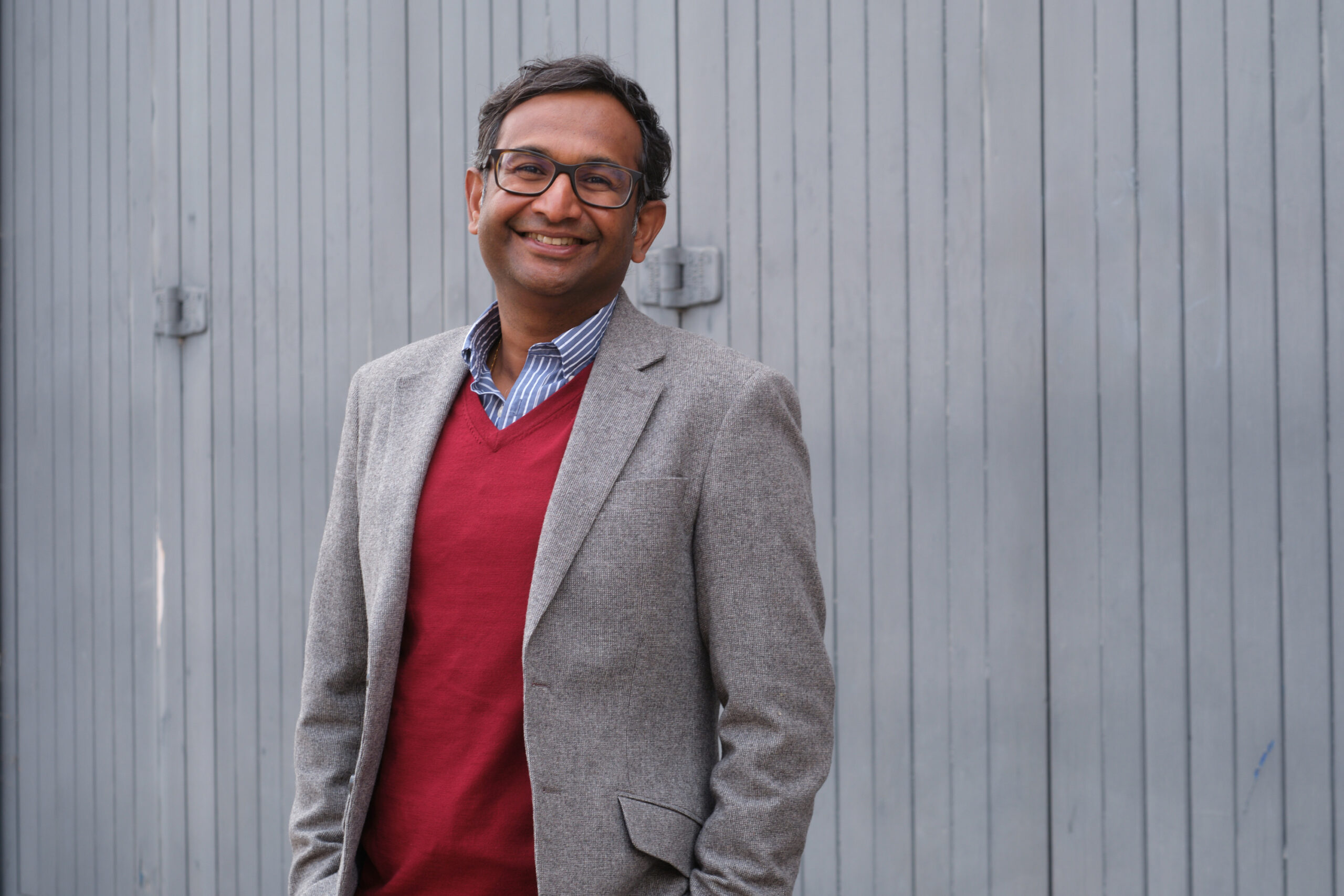 Romesh who is featured in the text wearing a red jumper and a grey jacket is standing in a relaxed manner against a grey slatted background.